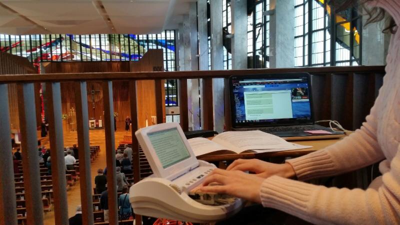 Image shows arms and hands of a woman using steno machine in the balcony of Lovers Lane church, with view of congregants and stained glass windows in the sanctuary below