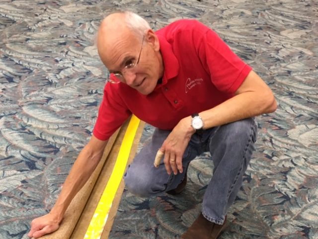 a man in a red shirt holds up a carpet, showing installation of the hearing loop line.