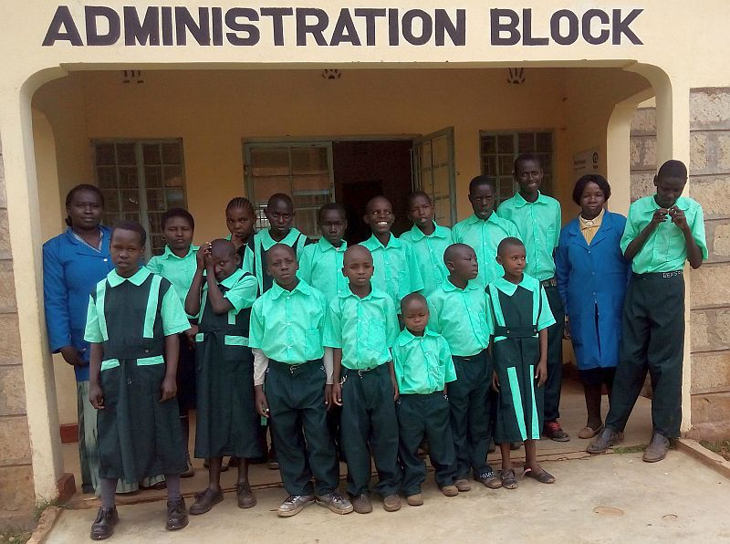 photo of students at the schook, they are standing in front of a building market Administration Block, there are 17 people in two rows, most are wearing green shirts and blue pants or skirts