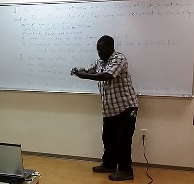 man gesturing, words of creed are written on a whiteboard behind him